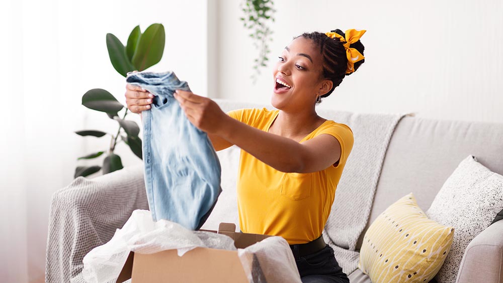 Delivery Concept. Smiling woman holding new jeans, unboxing cardboard package, sitting on the couch in living room at home, free space. Excited lady happy with purchase and clothes shipment